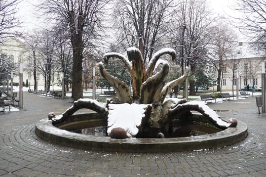 Sremska Mitrovica, Serbia 01.27.2023 City Fountain Stone flower in the central park. Snow cloudiness in winter. Snow covered city park. Decorative architectural forms. Trees, library and city archive