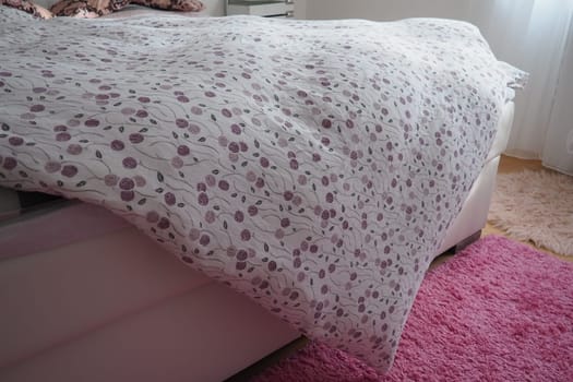 Cozy compact bedroom, white furniture, pink carpet and tulle curtains. Fluffy feather blanket thrown over the double bed. Romantic morning. Light from the window. The blanket hangs down to the floor