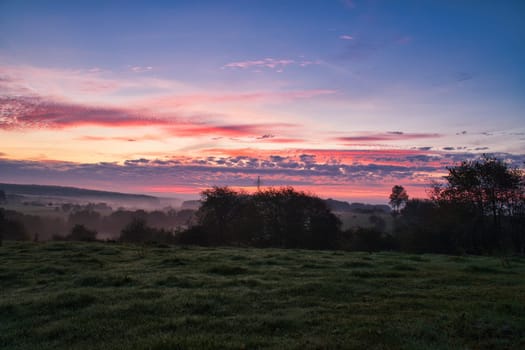 Sunrise over a neighboring forest with meadow in the foreground. Pasture landscape with tree standing alone at dawn. Landscape photo in Saarland