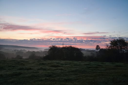 Sunrise over a neighboring forest with meadow in the foreground. Pasture landscape with tree standing alone at dawn. Landscape photo in Saarland