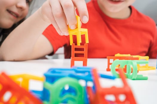 Happy little kids play toy colorful chairs sitting at table closeup. Portrait of boy and girl building from small plastic furniture. Home leisure, playtime. Entertaining interesting board game.