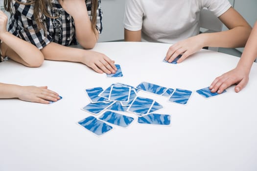 Group of friends play card game sitting at white table. Children take with hands flipped paper blue cards from pile. Interesting entertaining board game for kids. Home leisure, funny playtime.