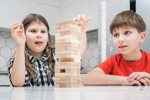 Cute school kids play jenga sitting at kitchen table. Boy and girl with concentrated facial expression build tower from wooden blocks. Home leisure, playtime. Entertaining interesting board game.