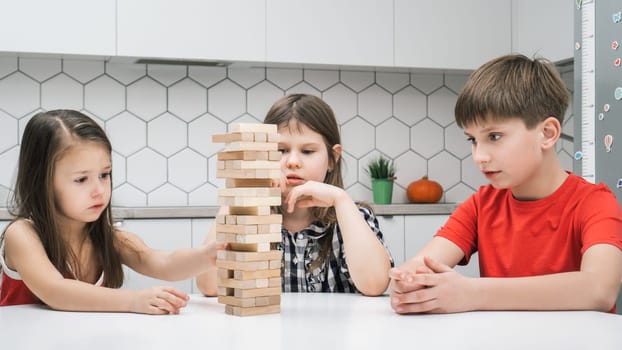 Concentrated school kids play tower sitting at kitchen table. Boy and girls build tower from little wooden blocks. Friends relax at home, playtime. Entertaining interesting board game.