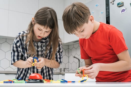 Cute school kids play toy constructor standing above kitchen table and looking down. Boy and girl collect plastic colored details of lego. Home leisure, playtime. Entertaining interesting board game.