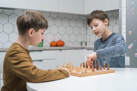 Cute schoolboys play chess sitting at kitchen table, side view. Wooden chessboard with black and white figures. Game strategy, tactics. Home leisure, playtime, hobby. Educational board game.