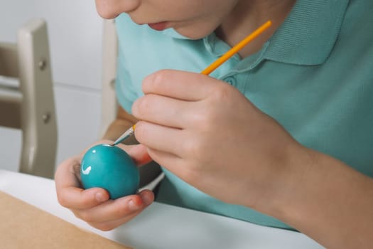 Cropped portrait of boy hand, painting multi coloring Easter hens eggs on brown paper plate table with paintbrush. Making art design white patterns on blue, red animal eggs. Holiday event. Close up