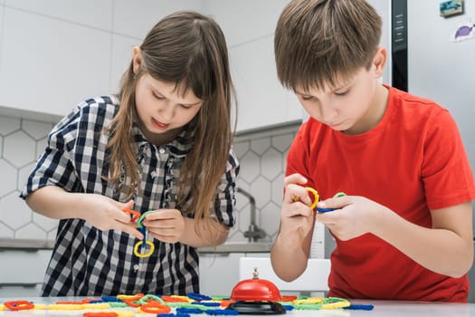 Funny school kids play toy constructor standing above kitchen table and looking down. Boy and girl collect plastic colored details of lego. Home leisure, playtime. Entertaining interesting board game.
