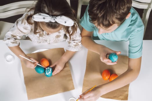 Portrait of two children, little girl and boy siblings, painting multi coloring Easter hens eggs in kitchen, with paintbrushes, sitting chair, paper table. Making art design white patterns. Top view.