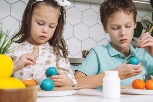 Portrait of studiously joyful two children, little boy and girl, painting brush multi coloring Easter hens eggs on paper plate table. Making white art patterns in kitchen. Traditional event. Close-up