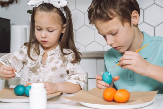 Portrait of enthusiastic two little children, boy and girl, painting brush multi coloring Easter hens eggs paper plate table, plant in kitchen. Making white art patterns. Traditional holiday event.
