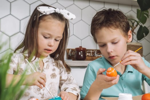 Portrait of concentrated joyful two children, little boy and girl, painting brush multi coloring Easter hens eggs on paper plate table. Making white art design patterns in kitchen. Light day. Close-up