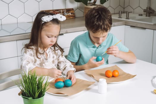 Portrait of studiously two little children, boy and girl, painting brush multi coloring Easter hens eggs on paper plate table, plant in kitchen. Making white art patterns. Traditional holiday event.