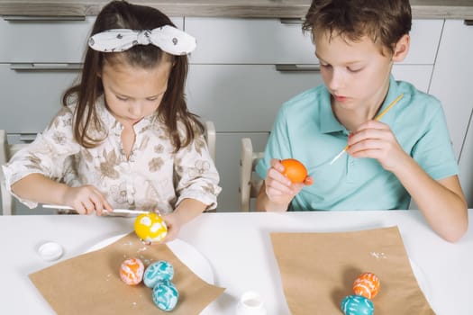 Portrait of two children, little girl and boy siblings, painting multi coloring Easter hens eggs in kitchen, with paintbrushes, sitting chair, paper table. Making art design white patterns. Top view.