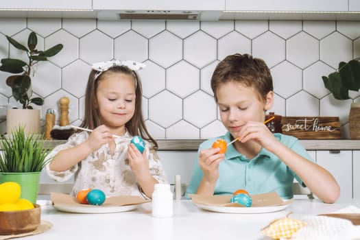 Portrait of concentrated smiling two kids, little girl and boy, coloring patterns on Easter hens eggs on paper table with paintbrushes near plant. Making art design. Sweet home nameplate, drawers.