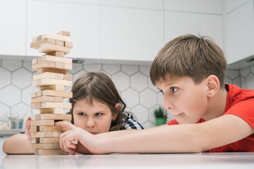 Happy school kids play tower sitting at kitchen table. Concentrated boy and girl build tower from little wooden blocks. Friends relax at home, playtime. Entertaining interesting board game.