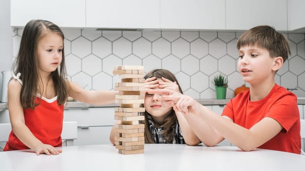 Happy school kids play tower sitting at kitchen table. Cute boy and girls build tower from little wooden blocks. Friends relax at home, playtime. Entertaining interesting board game.