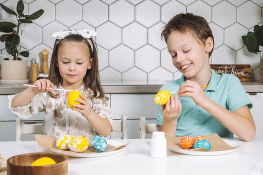 Portrait of concentrated joyful two children kids little girl and boy siblings, painting multi coloring Easter hens eggs in kitchen on paper table with paintbrushes. Making art design white patterns.