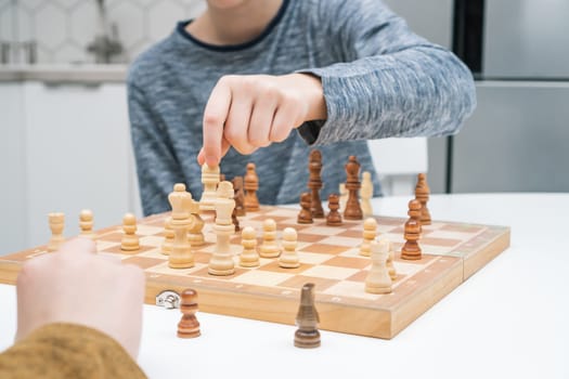 Clever boys play chess sitting at kitchen table. Hand of child move white figure on wooden chessboard. Game strategy, tactics. Home leisure, playtime, hobby. Educational board game.