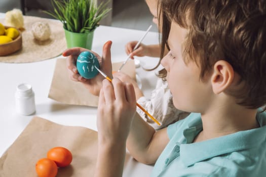 Cropped portrait of boy hand, painting multi coloring Easter hens eggs on brown paper plate table with paintbrush. Making art design white patterns on blue, red animal eggs. Holiday event. Close up