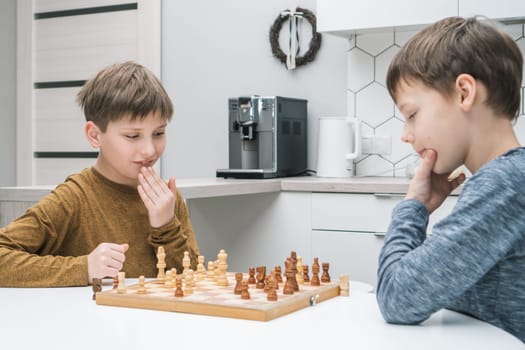 Thinking schoolboys play chess sitting at kitchen table, side view. Wooden chessboard with black and white figures. Game strategy, tactics. Home leisure, playtime, hobby. Educational board game.
