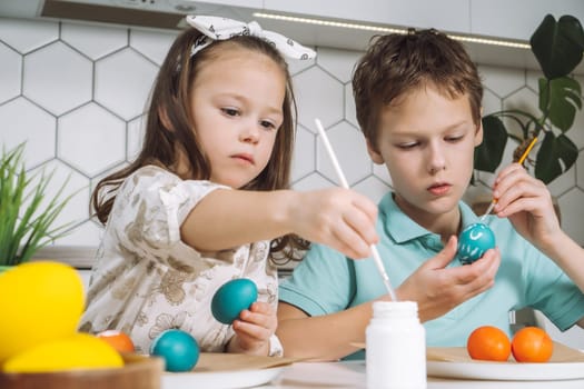 Portrait of concentrated joyful two children, little boy and girl, painting brush multi coloring Easter hens eggs on paper plate table. Making white art design patterns in kitchen. Light day. Close-up