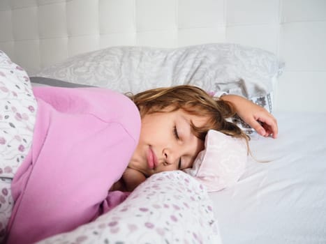 A beautiful caucasian girl of 8 years old with blond hair, dressed in pink pajamas, sleeps on a bed with a fluffy blanket, hugging a pillow. Soft light of the morning sun streams through the window