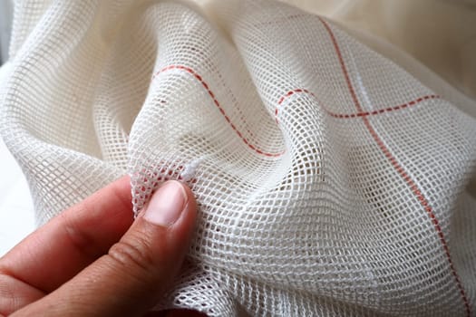 White coarse cotton or linen curtains made of weaves of thin threads. A woman's hand is feeling the fabric. Window decoration room. Red stripe. The fabric is folded in soft waves. Apartment interior