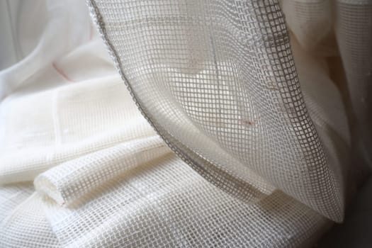 White coarse cotton or linen curtains made of weaves of thin threads. Simple compact economical apartment interior. Window decoration in the room. Red stripe. The fabric is folded in soft waves.