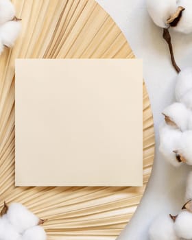 Blank card on dried palm leaf with cotton flowers top view, mockup. Romantic scene with square paper card. Bohemian pastel greeting or wedding Invitation .