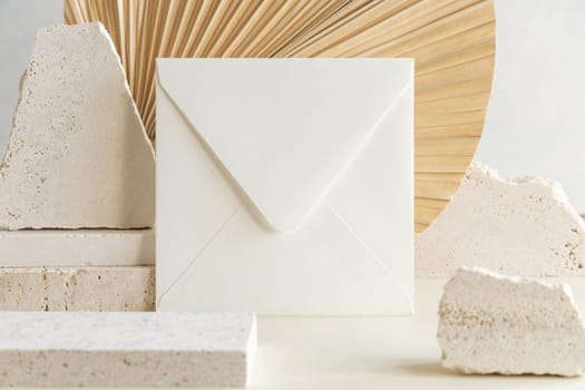 Blank square envelope near beige travertine stones against dry beige palm leaf close up, mockup. Boho scene with horizontal paper card. Bohemian greeting or wedding Invitation