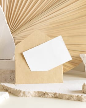 Blank card and envelope near beige travertine stones against dry beige palm leaf close up, mockup. Boho scene with horizontal paper card. Bohemian greeting or wedding Invitation