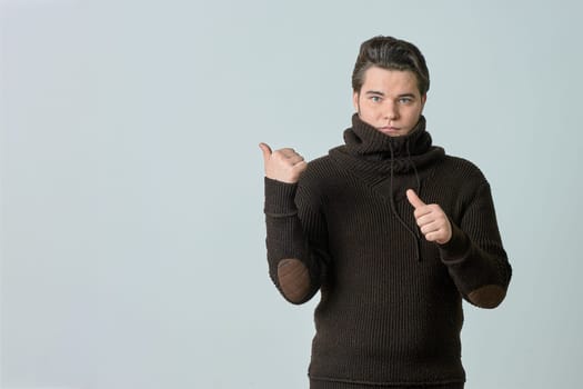 a man in a brown sweater and a bright color points to something and perfectly, showing an empty space with his thumb, shows everything on a light background, copy space. High quality photo