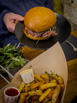 A person holds a freshly prepared burger topped with greens, accompanied by a generous serving of crispy fries and dipping sauces, creating a satisfying meal experience.
