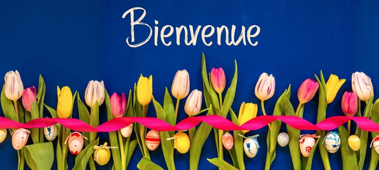 French Text Bienvenue Means Welcome. Banner Of White And Pink Tulip Spring Flowers With Ribbon And Easter Egg Decoration. Blue Wooden Background
