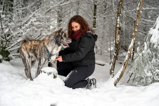 Young woman is kneeling next to her akita inu dog with gray fur, looking at her dog and smiling, in the forest during winter with lots of snow
