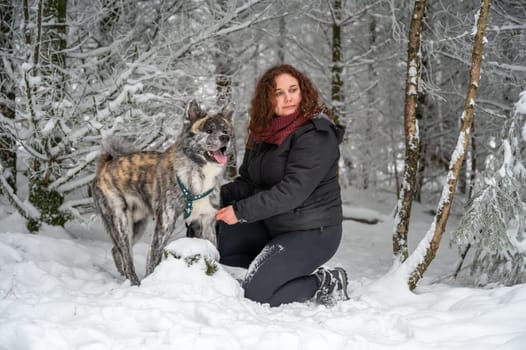 Young woman is kneeling next to her akita inu dog with gray fur, in the forest during winter with lots of snow