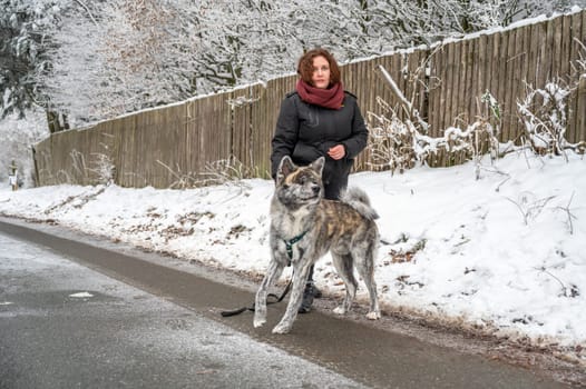 Young woman with brown curly hair standing on a frozen road with her gray colored akita inu dog, looking at camera during winter with lots of snow