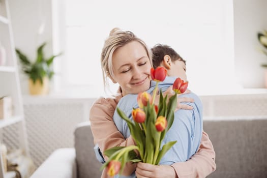 Cute boy sitting on the sofa with mom and giving a bouquet of tulips to her. Concept of the Mother's day and family relation.