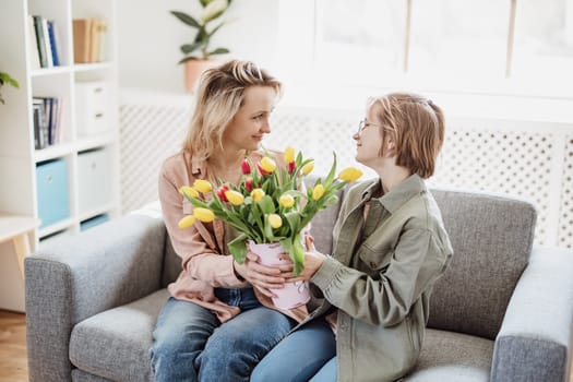 Mother and daughter sitting on the sofa with tulips in their hands indoor. Concept of the mother's day, birthday and motherhood.