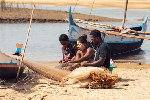Morondava, Madagascar - November 3, 2022: Fisherman and woman repairing fishing nets at the estuaries of a river, canal Hellot. The woman has a traditionally Malagasy painted face