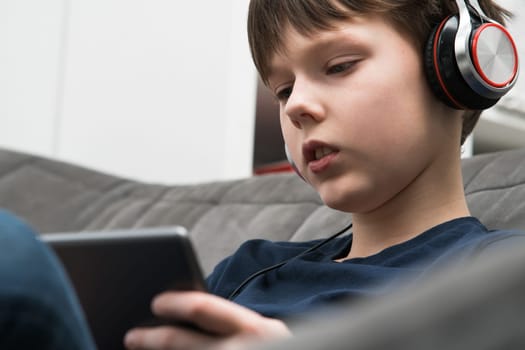 Calm, concentrated little boy in headphones watching movies and listen to music with digital tablet, pad in hands, sitting on sofa. Audiobooks, podcasts in your earphones. Portable wireless device