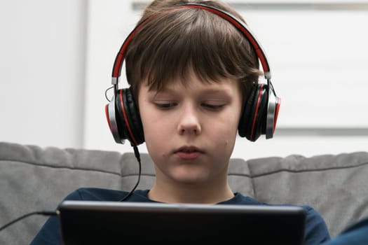 Passionate, concentrated brunette boy in wired headphones watching movies, listen to music with digital pad in hands, sitting on sofa. Portable gadget, musical equipment, sound quality and play online