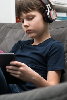 Vertical tranquil, concentrated little boy in headphones listen to music and watching movies with digital tablet, pad in hands, sitting on sofa. Portable Bluetooth device. Podcasts in your earphones.
