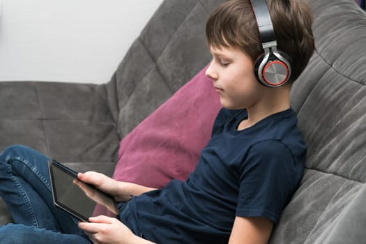 Joyful, spiritual little boy in headphones watching movies and listen to music with digital tablet and streaming service, sit on gray sofa. Audiobooks, podcasts in pad. Wireless gadget. Profile view