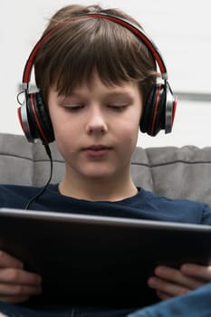 Vertical smiling, brunette boy in wired headphones watching movies and listen to music with digital tablet, pad in hands, sitting on sofa. DJ and best quality stereo sound, electronic equipment device