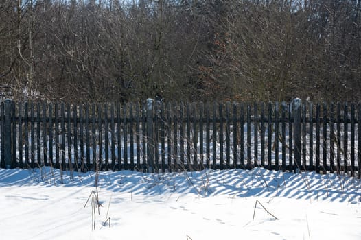An old wooden fence with snow in the sunlight with trees