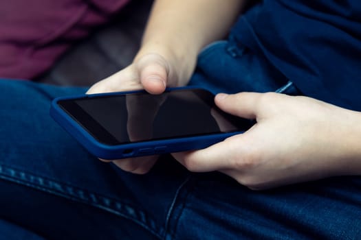 Close up unrecognizable teenage boy using smartphone with black screen in blue mobile case, sit on sofa. CEO, IT Internet technology, social media and smart applications. Investment and finance online