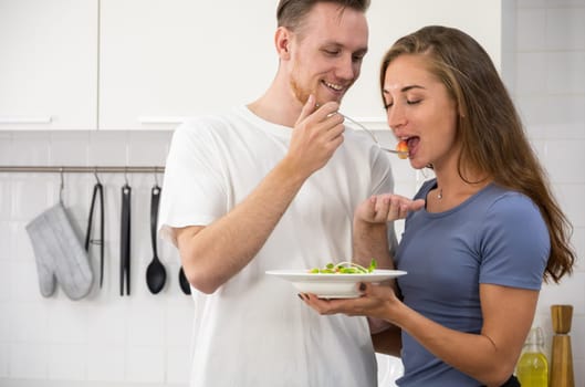 Smiling young couple in love making healthy vegan salad in kitchen, girl testing it from man hand. Happy family spending time together in weekend