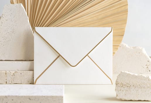 Blank envelope near beige travertine stones against dry beige palm leaf close up, mockup. Boho scene with horizontal paper card. Bohemian greeting or wedding Invitation